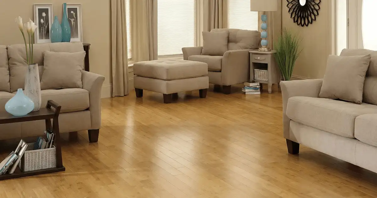 A living room with professionally installed bamboo flooring, beige sofas, a matching ottoman, a side table with a blue lamp, vases, framed pictures, and large windows with blinds and curtains in Grand Rapids, MI